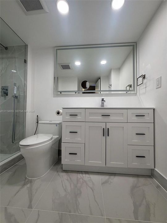 White vanity with multiple drawers and cabinets featuring matte black hardware, large-format tile flooring, frameless glass walk-in shower with stone-finish surround, and expansive wall-mounted mirror