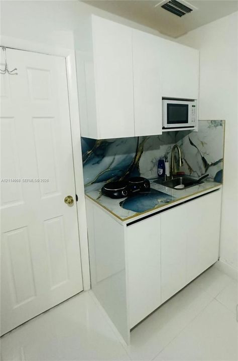 Compact kitchenette featuring white flat-panel cabinetry, patterned stone-style backsplash, integrated microwave, drop-in sink with a high-arch faucet, and polished flooring