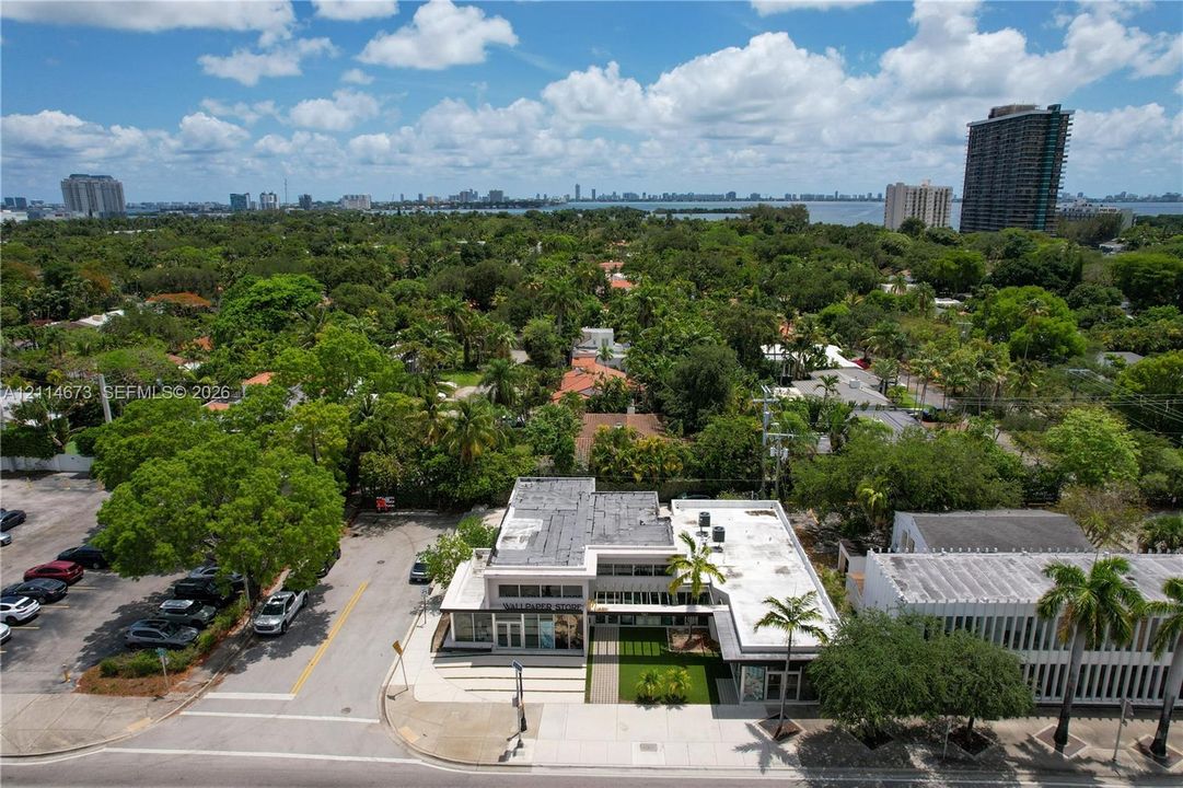 Aerial perspective of a multi-level commercial structure featuring a flat roofline, extensive exterior glazing, a paved corner lot, and integrated landscaping with mature palm trees