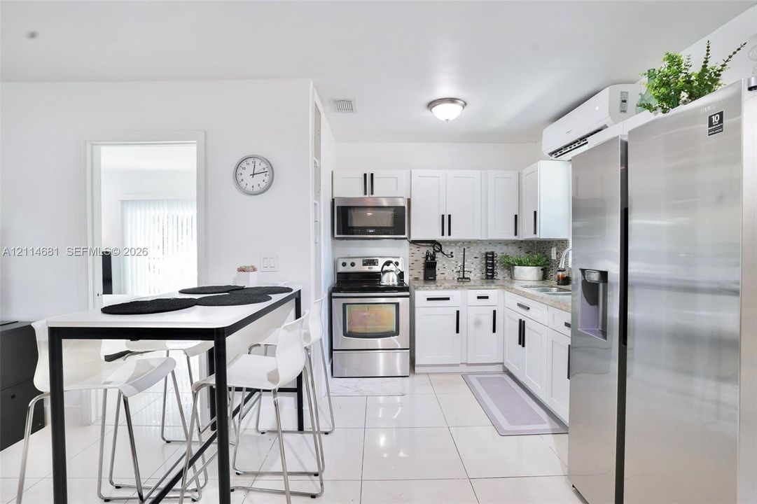 Kitchen featuring white shaker-style cabinetry, stainless steel appliances, patterned tile backsplash, neutral stone-finish countertops, and white tile flooring