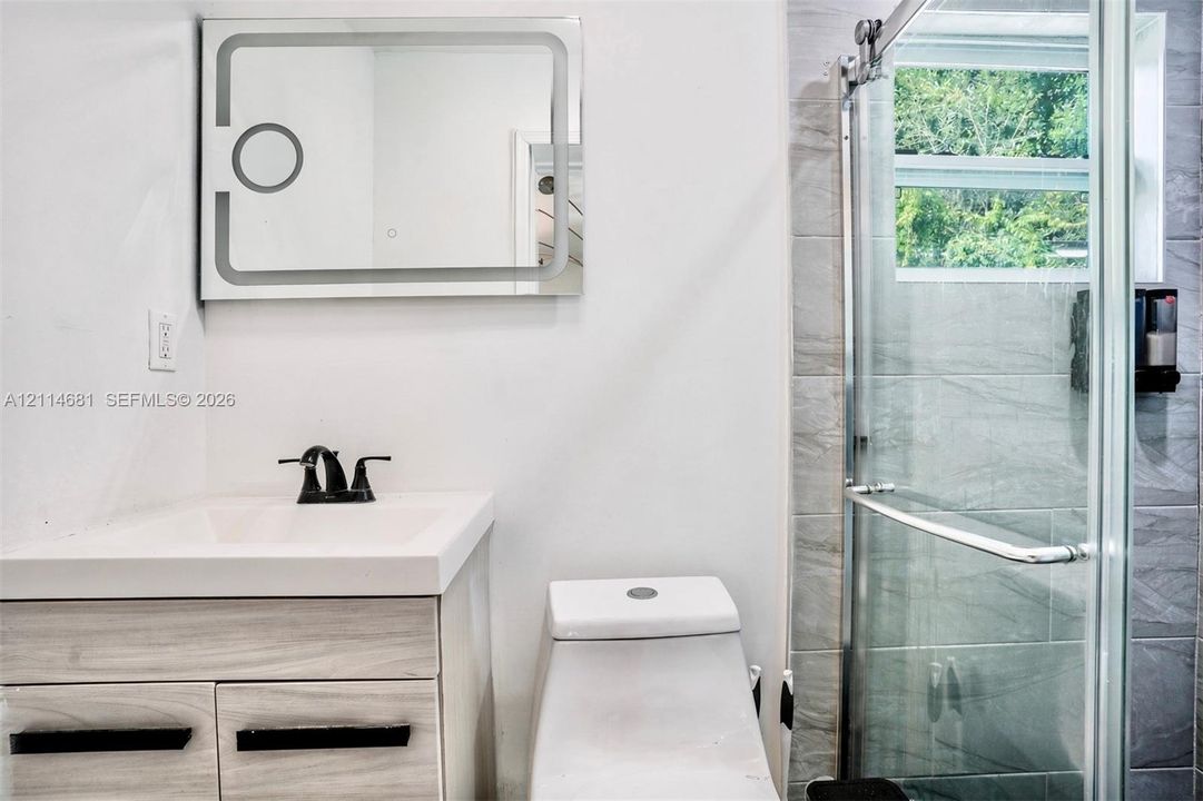 Wood-finish vanity with integrated sink, matte black faucet, illuminated mirror, and walk-in shower featuring a sliding glass enclosure and textured wall tiling