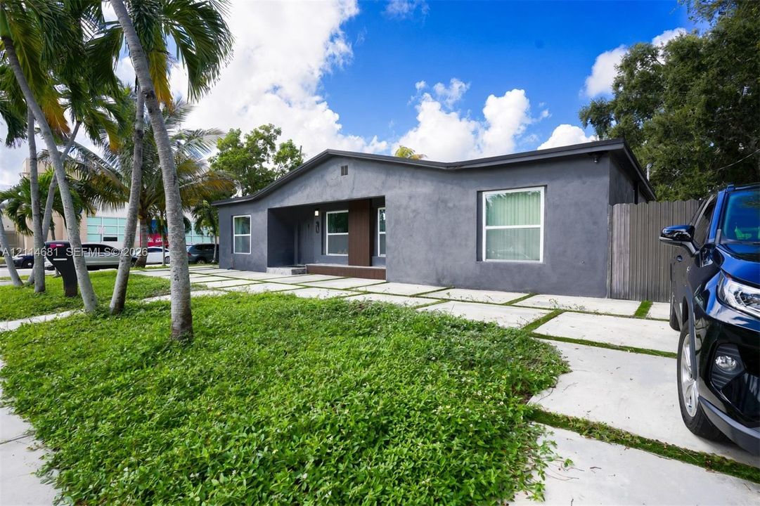 Single-story stucco exterior featuring a paved concrete grid driveway, recessed entryway with wood-tone accents, and mature palm trees