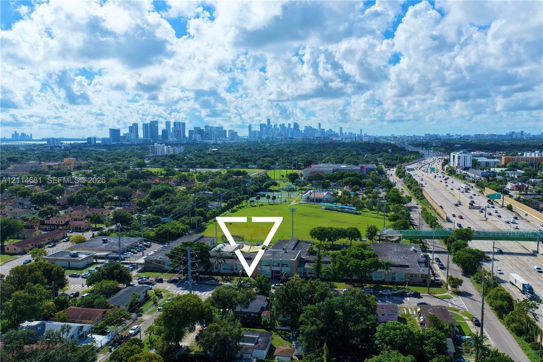 Aerial perspective of the property grounds featuring a multi-story building, established mature trees, proximity to expansive green space, and a backdrop of the urban skyline