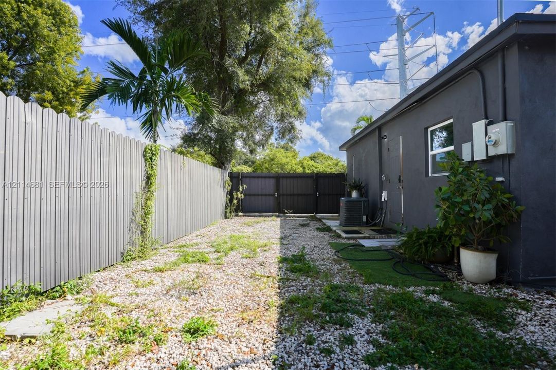 Side yard enclosed by corrugated metal fencing, featuring a gravel surface, palm tree, and gated entry