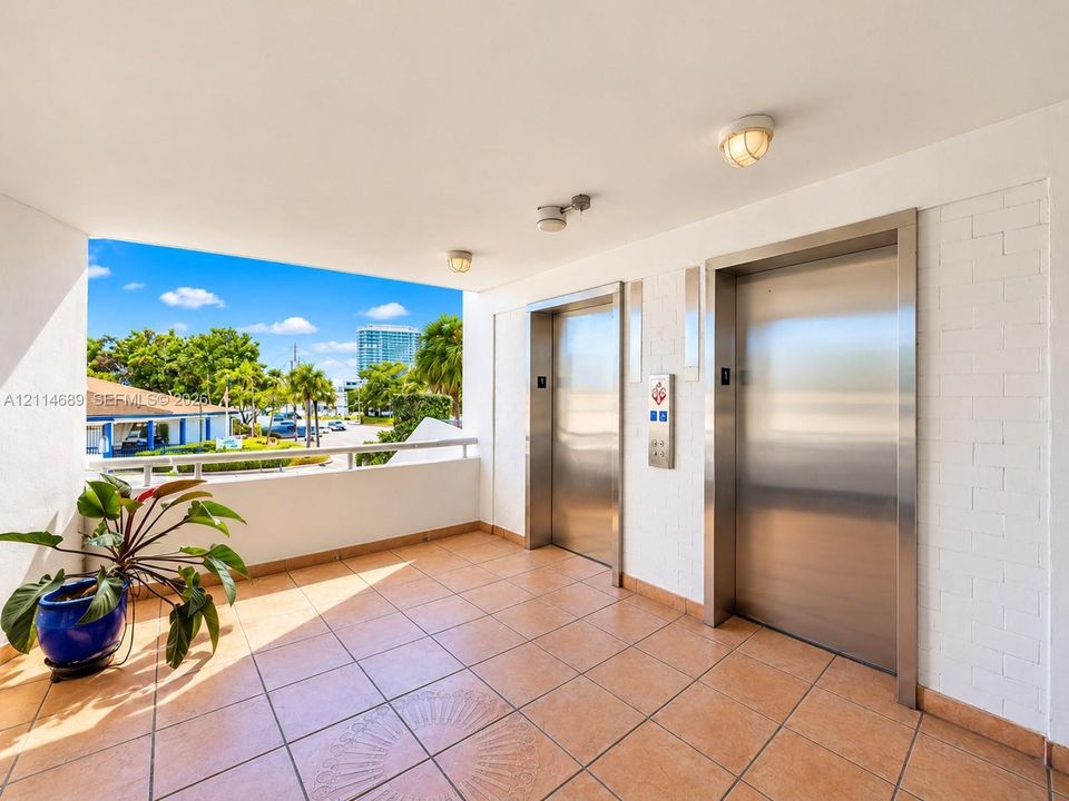 Elevator lobby featuring tiled flooring, dual stainless steel elevator doors, white painted brick accent wall, and perimeter metal railing