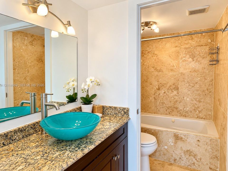 Bathroom featuring a vanity with speckled stone countertop, vessel sink with a metal faucet, large wall mirror, and a tub-shower surround finished with oversized stone-tile wall cladding