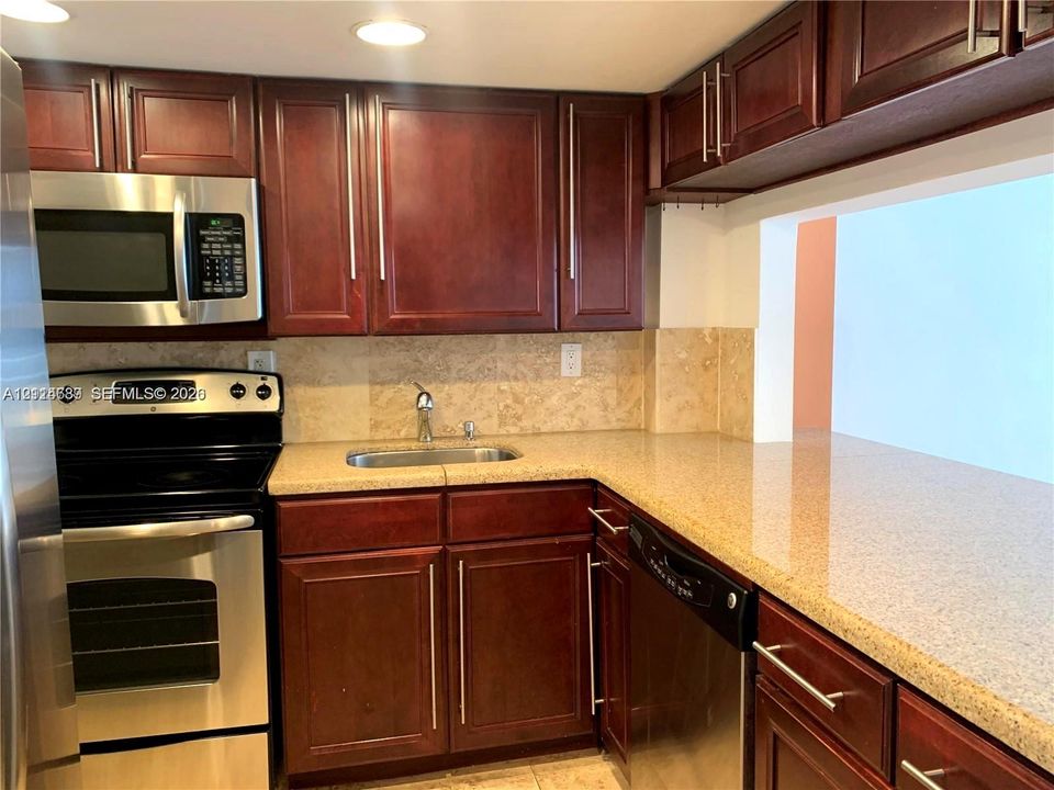 Kitchen featuring rich-toned cabinetry with long-pull hardware, solid-surface countertops, stone-tile backsplash, and stainless-steel appliances