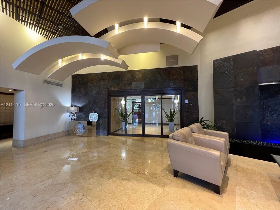Grand lobby entrance featuring tiered, curved architectural ceiling elements with recessed lighting, large-format stone floor tiles, and a prominent dark natural stone accent wall