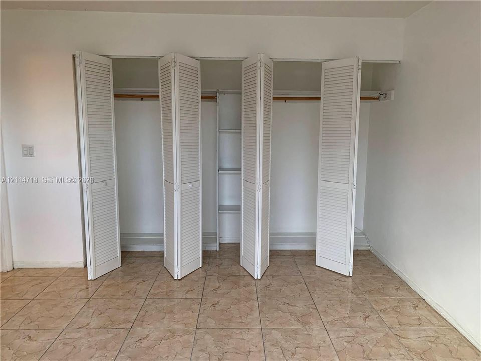 Large closet featuring white bifold louvered doors, integrated shelving, and horizontal hanging rods, complemented by neutral tile flooring