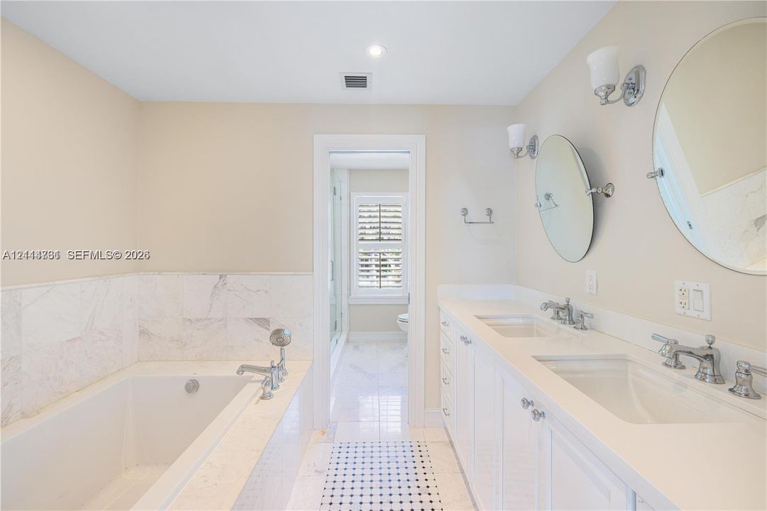 Double vanity with integrated sinks, white cabinetry, polished metal fixtures, and wall-mounted mirrors, featuring a soaking tub with stone tile surround and stone flooring