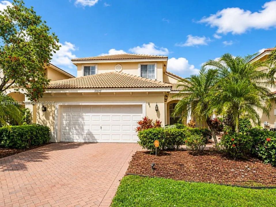 Stucco exterior with a tiled roof, paver driveway, two-car garage, and landscaped grounds featuring palm trees