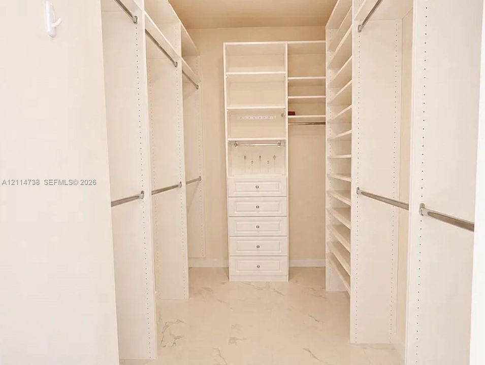 Walk-in closet featuring white built-in shelving, multiple hanging rods, a central storage tower with five soft-close drawers, and neutral stone-pattern flooring