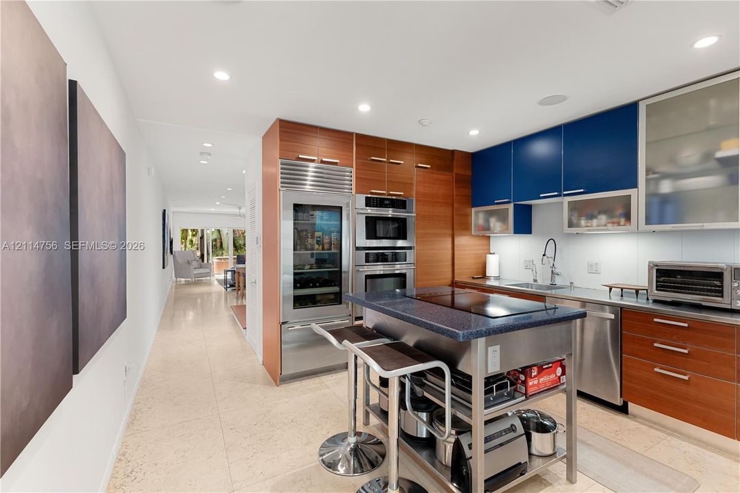 Kitchen featuring custom wood-veneer and navy cabinetry, professional-grade stainless steel appliances, stone-topped island with an integrated cooktop, recessed lighting, and stone flooring