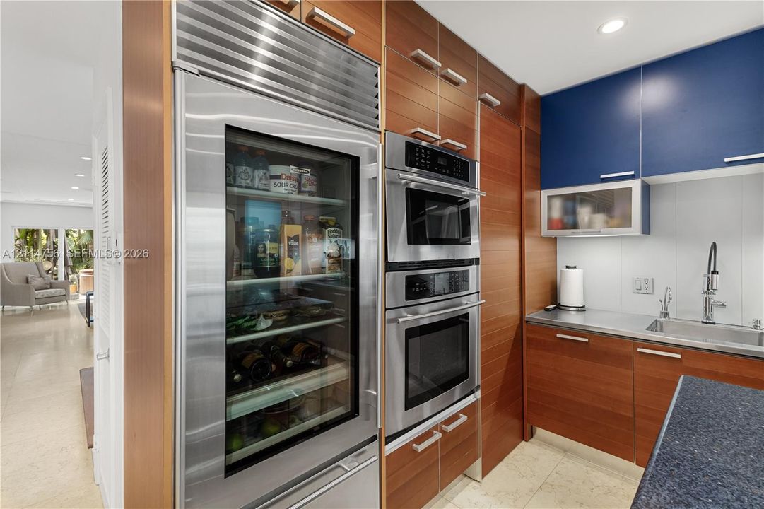 Built-in stainless steel refrigerator with glass door, dual wall-mounted ovens, custom wood-veneer cabinetry, blue upper cabinets, and a stainless steel sink with high-arc faucet