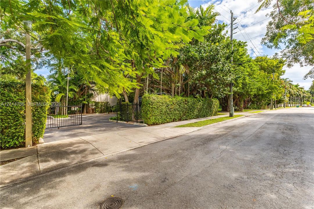 Exterior setting featuring a gated asphalt entrance, mature perimeter landscaping, concrete sidewalk, and established canopy trees