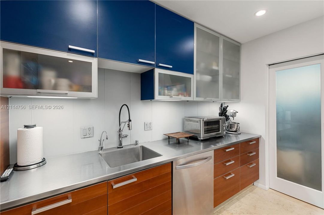 Modern kitchen featuring dual-tone cabinetry with blue upper units and wood-finish lower drawers, metallic countertops, integrated stainless steel dishwasher, and frosted glass door