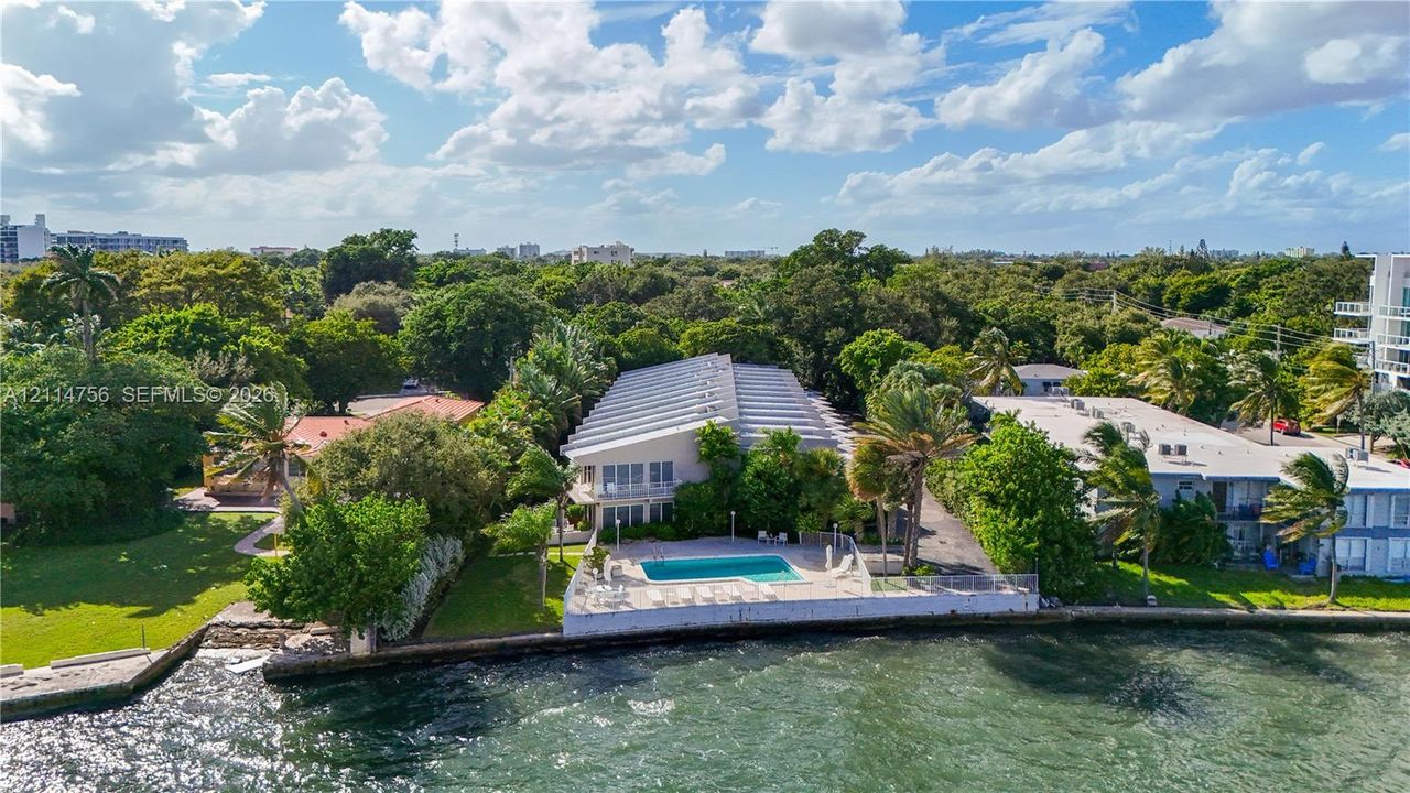 Waterfront property featuring a concrete seawall, in-ground swimming pool, paved patio, and mature landscaping