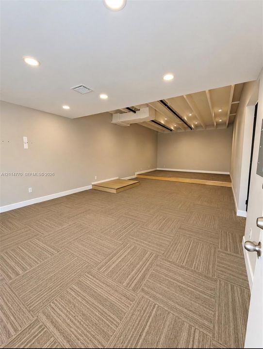 Open-concept interior featuring recessed lighting, patterned carpeting, exposed ceiling beams, and a tiered platform
