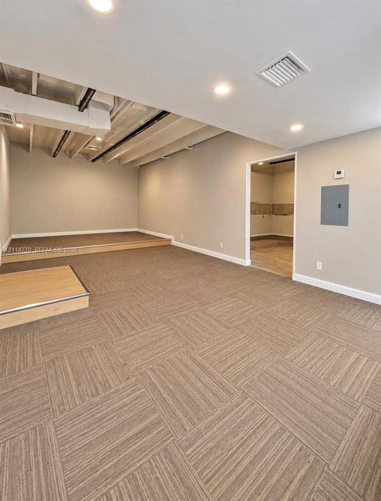 Interior space featuring square-patterned carpet tiles, tiered raised platforms, recessed ceiling lighting, and an exposed structural ceiling