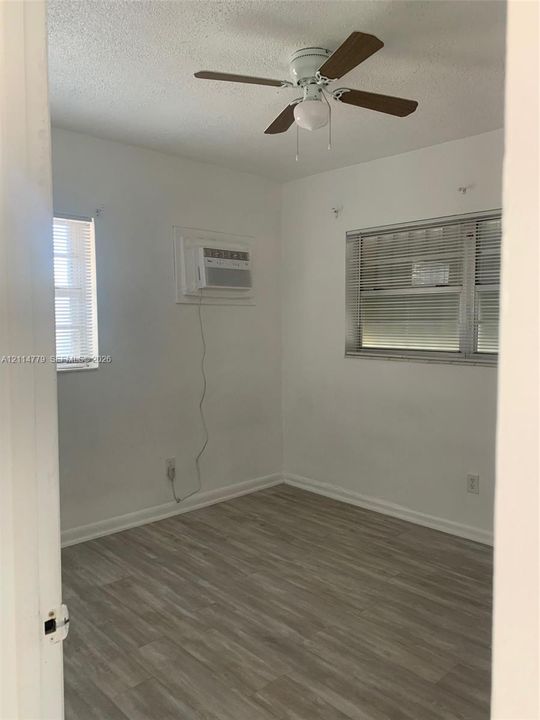 Room featuring wood-finish flooring, a ceiling fan with integrated lighting, two windows with horizontal blinds, and a built-in wall-mounted air conditioning unit