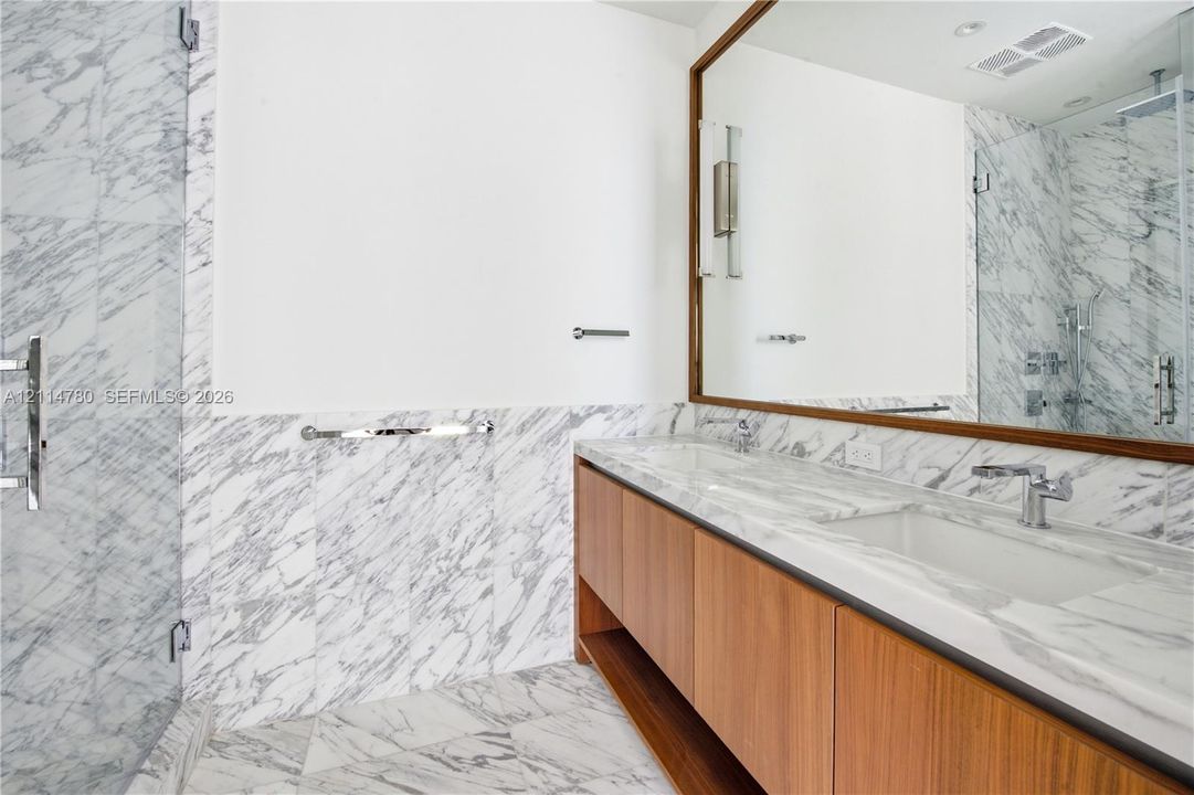 Floating wood-finish vanity with integrated dual sinks, stone countertops, full-wall vanity mirror, and glass-enclosed shower featuring floor-to-ceiling stone wall tiling