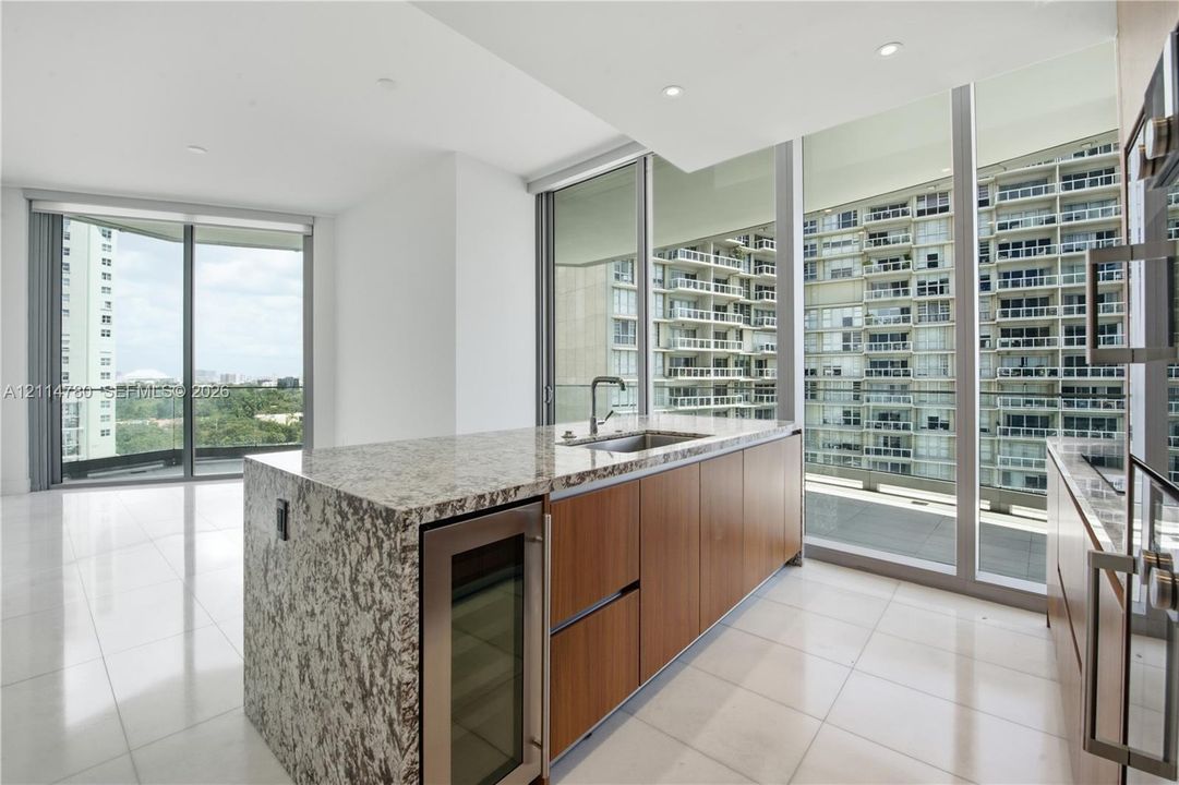 Kitchen island featuring a stone-finish countertop, wood-finish cabinetry, a built-in wine refrigerator, stainless steel undermount sink, and floor-to-ceiling glass windows
