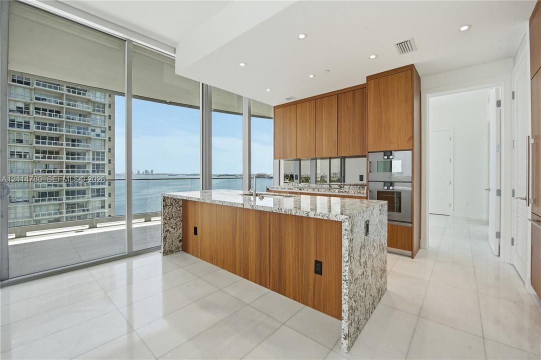 Modern kitchen featuring wood cabinetry, a waterfall-edge stone island, built-in stainless steel double ovens, and floor-to-ceiling glass paneling with water and urban scenery