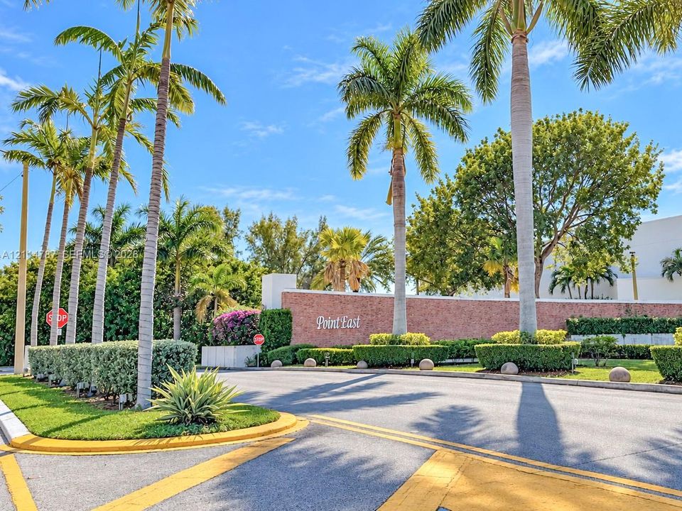 Entrance featuring a brick monument wall, mature palm trees, manicured hedging, and asphalt drive