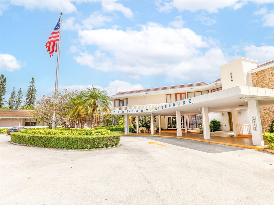 Clubhouse exterior featuring a covered porte-cochère, stone masonry facade, manicured landscaping, and asphalt parking area