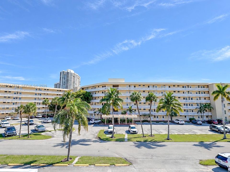 Multi-story residential complex featuring continuous exterior balcony railings, a central covered porte-cochère entry, surface parking, and mature palm tree landscaping