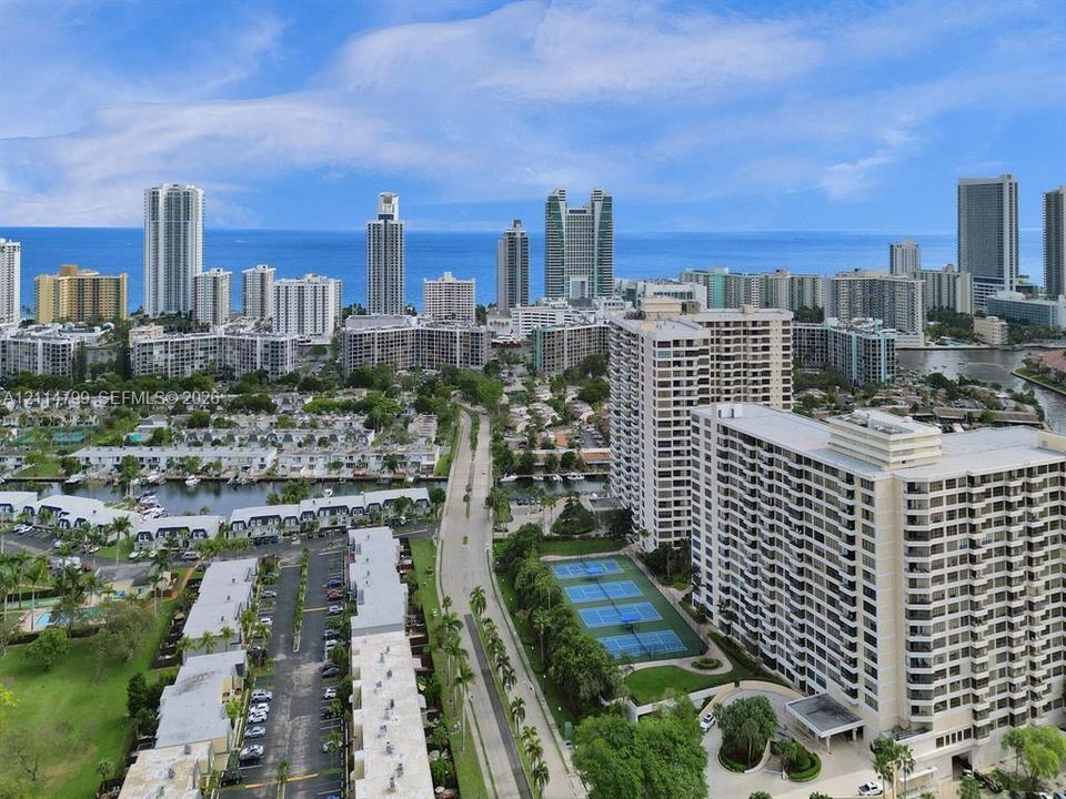 Aerial perspective featuring coastal urban surroundings, a multi-story high-rise complex, tennis courts, and mature landscaping