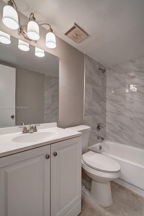 Bathroom featuring a vanity with a white countertop and integrated sink, full-width mirror, polished chrome lighting fixture, and a tub-shower combination with floor-to-ceiling stone-finish tile surround
