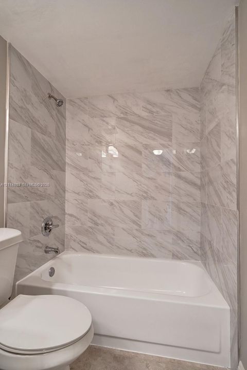 Bathtub with a white acrylic surround, large-format grey-toned veined tile wall cladding, wall-mounted chrome shower head, and chrome plumbing fixtures