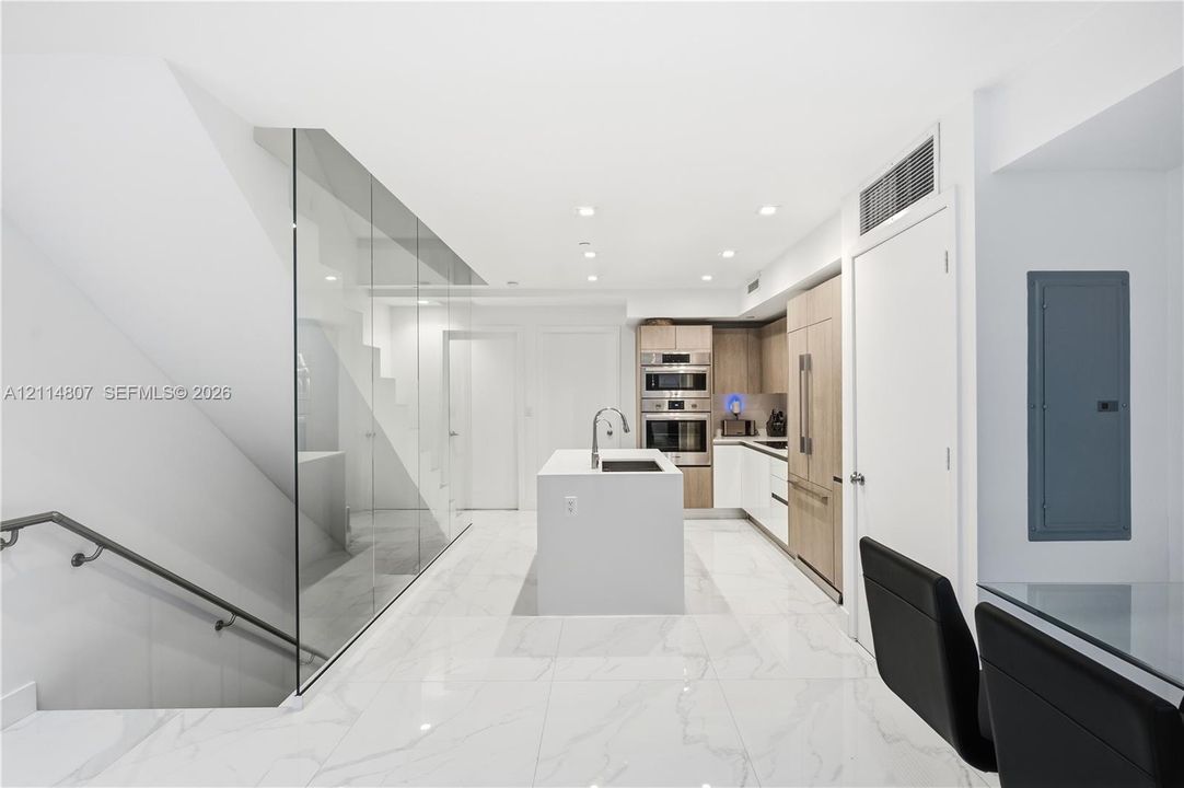 Modern kitchen featuring light wood-finish cabinetry, built-in stainless steel appliances, a central island with a sink, and stone-finish tile flooring