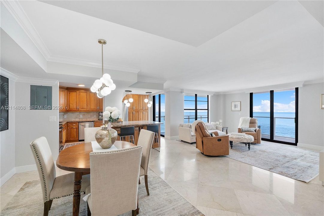 Open-concept interior featuring tray ceilings, natural stone flooring, floor-to-ceiling windows with water backdrop, and custom cabinetry with integrated lighting