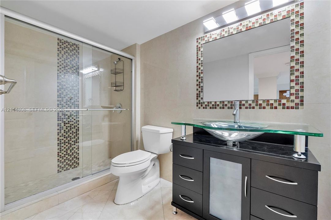 Modern vanity featuring a black finish, glass countertop, and vessel sink, paired with a glass-enclosed shower with mosaic tile accents, matching mosaic tile framed mirror, and light-toned tile flooring