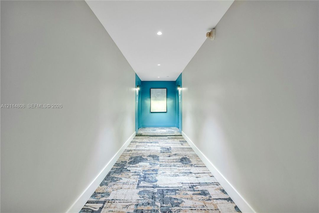Hallway featuring neutral-toned walls, patterned textile floor covering, recessed ceiling lighting, and wall-mounted light fixtures