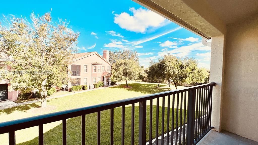 Covered balcony featuring a black metal railing, overlooking a landscaped lawn and mature trees