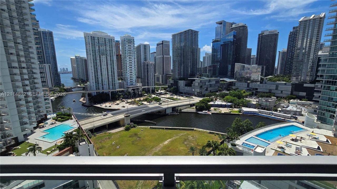High-rise balcony featuring metal railing, panoramic cityscape, waterfront exposure, and two outdoor swimming pools