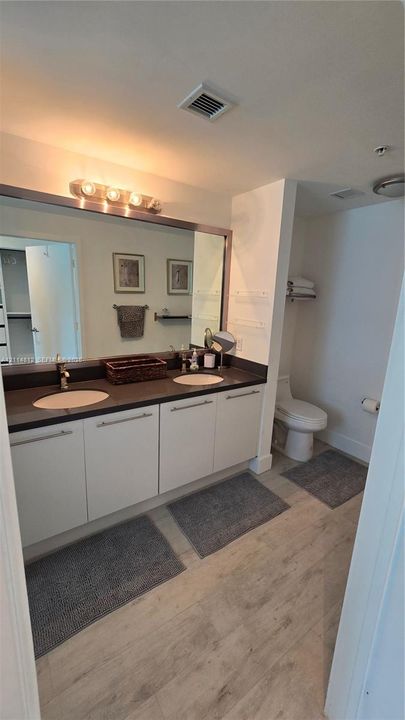 Dual-sink vanity with a dark solid surface countertop, undermount basins, wood-finish flooring, and a full-width vanity mirror with integrated lighting