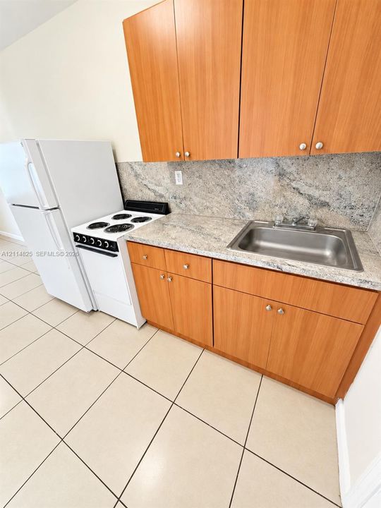 Kitchenette featuring wood-finish cabinetry, stone-surface countertops, matching stone backsplash, stainless steel drop-in sink, and ceramic tile flooring