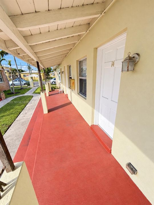 Covered walkway featuring a painted concrete porch, exposed beam ceiling, multiple exterior entry doors, and wall-mounted metal fixtures