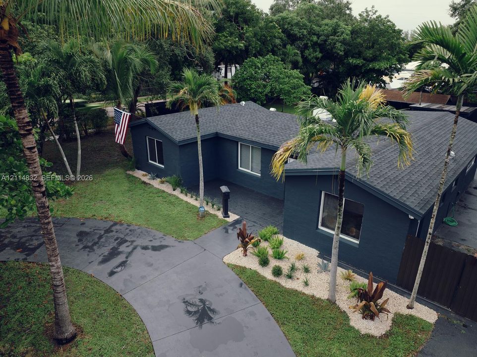 Single-story exterior featuring dark-toned masonry siding, a shingled roof, curved paved driveway, and established palm tree landscaping with decorative gravel beds