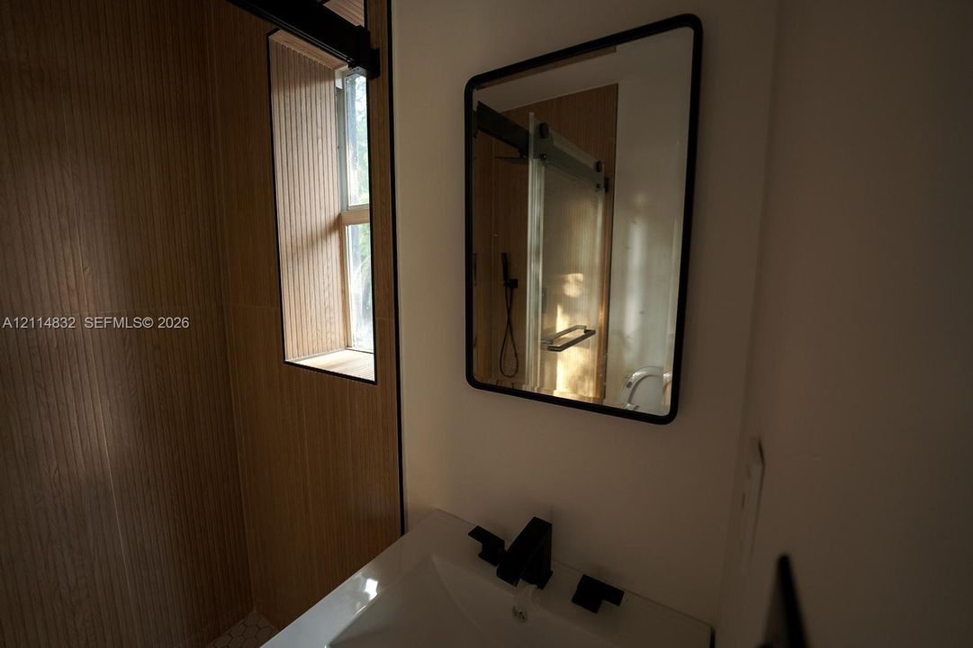 Bathroom featuring a white rectangular sink with matte black widespread faucet, black-framed mirror, wood-finish wall paneling, and glass-enclosed shower with black hardware