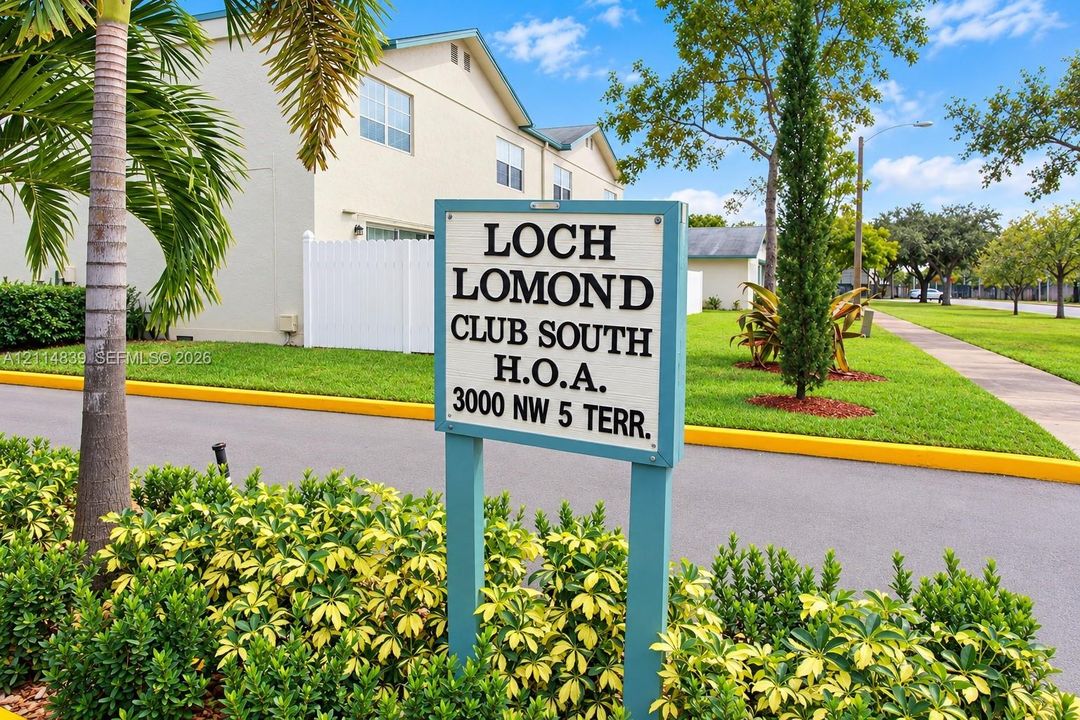 Exterior featuring a light-toned stucco facade, white vinyl fencing, paved asphalt pathways, and mature palm and cypress landscaping