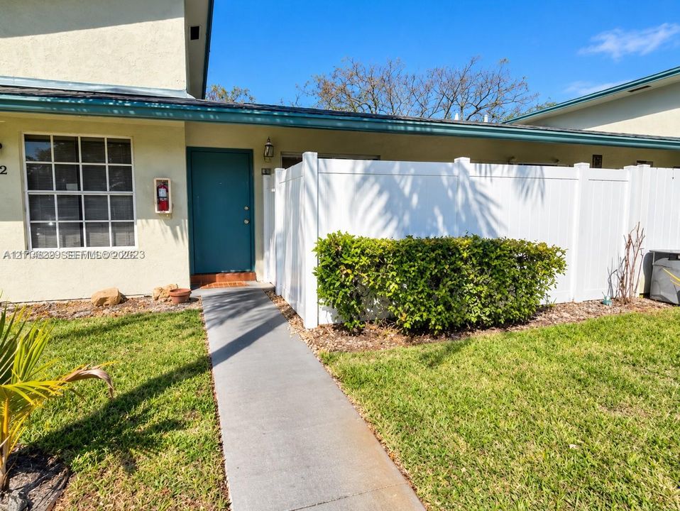 Exterior entrance featuring a solid door, concrete walkway, white privacy fencing, and maintained shrubbery