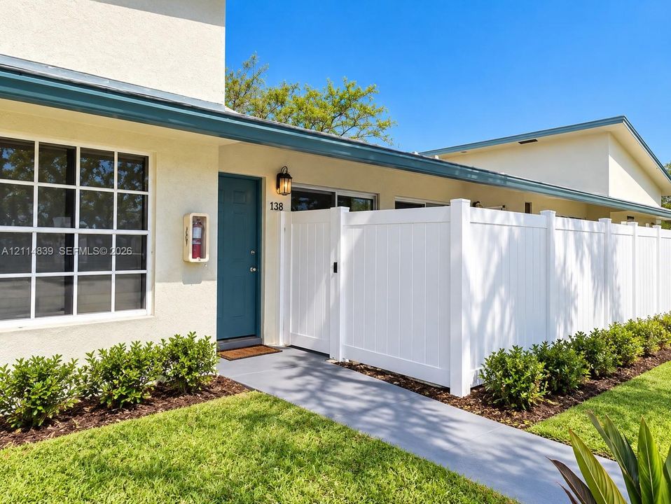 Exterior facade featuring smooth stucco finish, grid-pane windows, covered entryway with wall-mounted lighting, and a white vinyl privacy fence