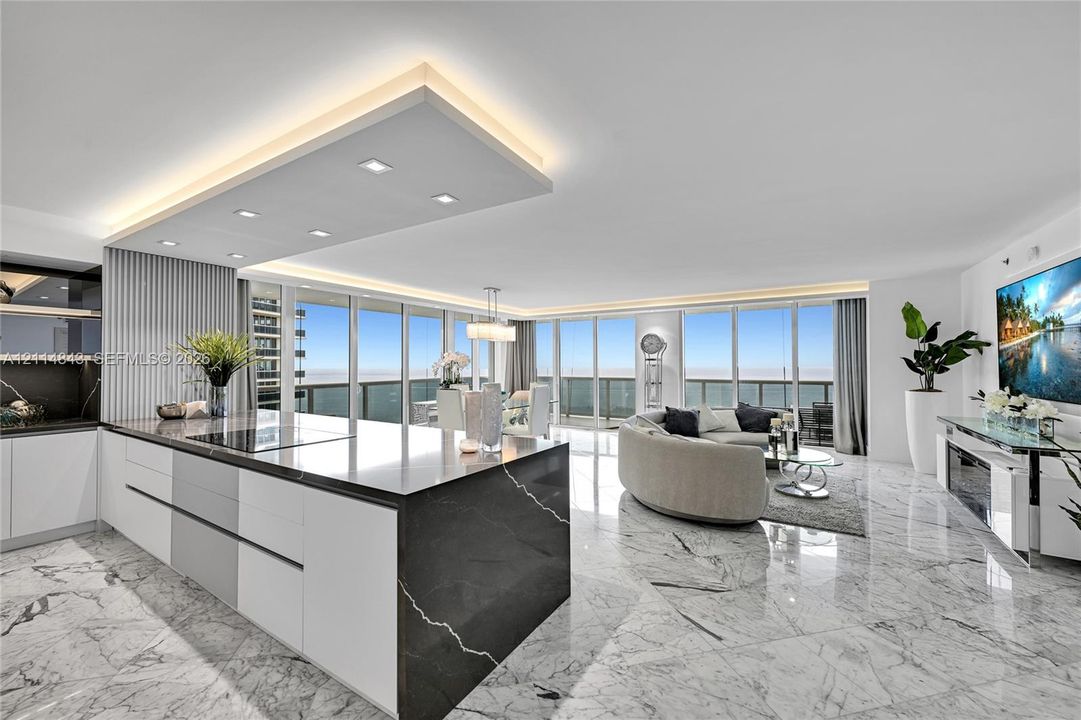 Open-concept living space featuring stone flooring, floor-to-ceiling windows, a center kitchen island with a dark stone waterfall countertop, recessed ceiling lighting, and custom cabinetry