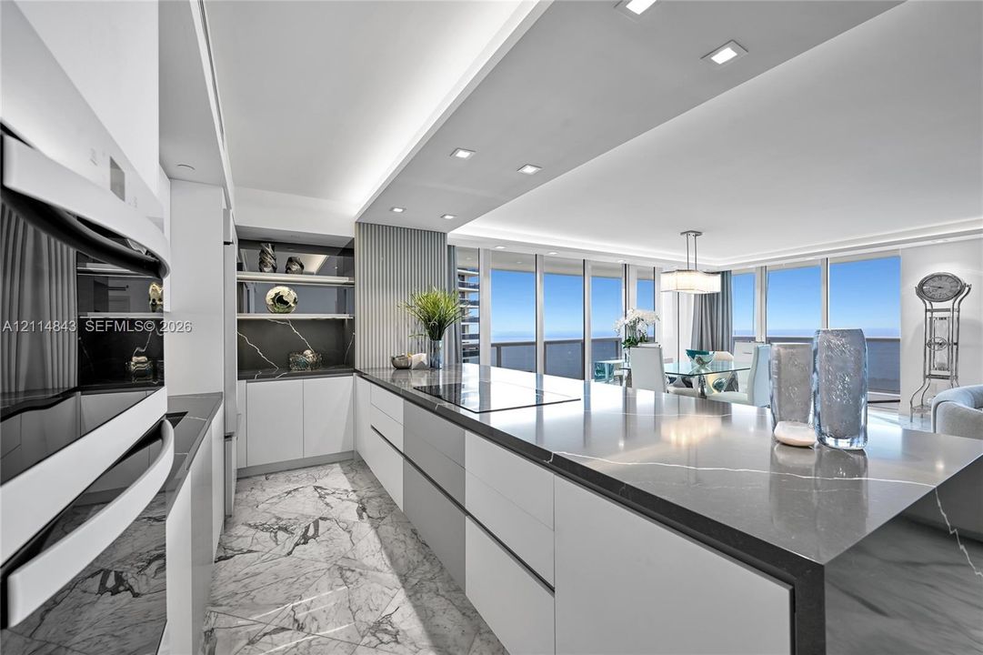Contemporary kitchen featuring custom flat-panel cabinetry, a large waterfall-edge island with solid surface countertop, built-in appliances, recessed lighting, and stone-finish flooring