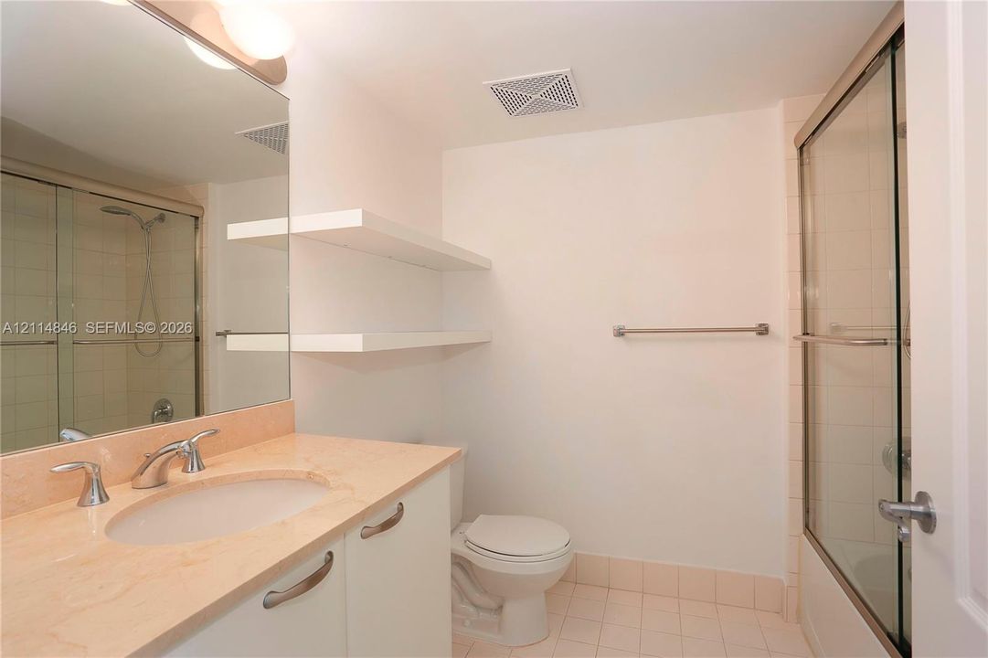 Bathroom featuring a vanity with a neutral-toned countertop and integrated sink, large wall-mounted mirror, white built-in shelving, and a glass-enclosed shower with tiled surround
